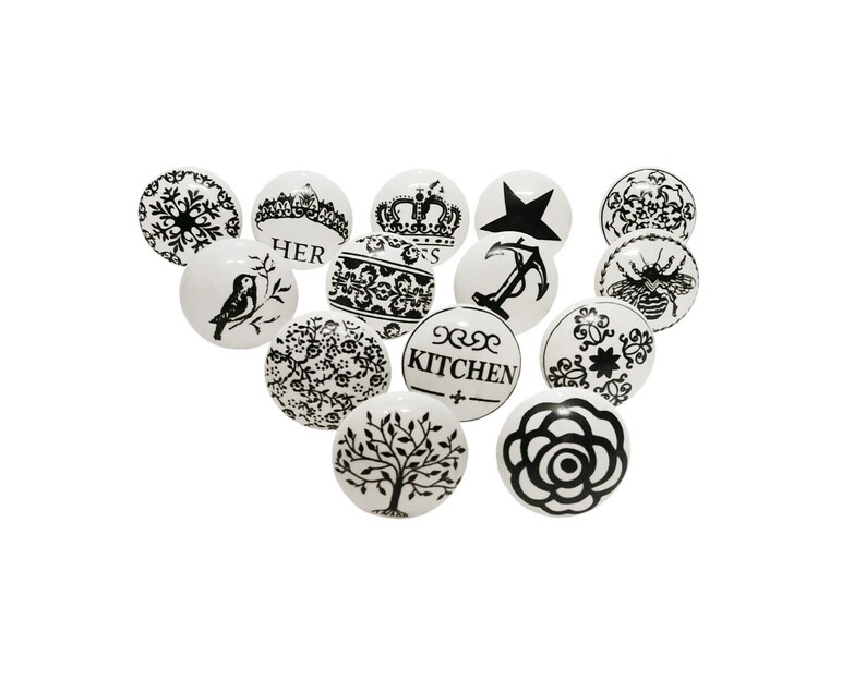 May include: Set of 12 white ceramic cabinet knobs with black designs. The designs include a snowflake, a crown, an anchor, a bird, a bee, a rose, a tree, and various floral patterns. Some knobs have the words "HER" and "KITCHEN" printed on them.