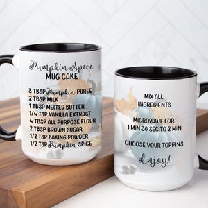 May include: Two white ceramic mugs with black handles and rims, featuring a pumpkin spice mug cake recipe. The mugs are decorated with watercolor pumpkins and fall leaves. The mugs are on a wooden cutting board.