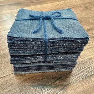 May include: A stack of denim fabric squares, tightly bound with blue twine. The denim has a classic blue jean color and a textured appearance. The stack is neatly arranged, suggesting a craft or sewing project.