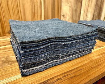 May include: A stack of blue denim fabric squares, neatly arranged on a wooden surface. The edges of the fabric are frayed, and the denim has a slightly worn appearance. The squares are stacked in a rectangular shape.