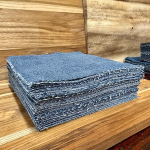 May include: Stacks of square, blue denim fabric pieces with frayed edges. The fabric is a medium wash denim, and the stacks are neatly arranged on a wooden surface. The image shows a close-up of the fabric, highlighting the texture and color.