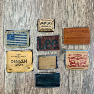May include: A collection of nine leather and fabric patches from various clothing brands, including Levi Strauss & Co., Lee, and Lucky Brand. The patches feature brand names, logos, and text, such as "American Eagle" and "Proud to Protect."