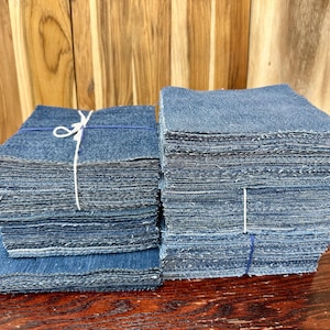 May include: Two stacks of denim fabric squares, each bound with string. The denim is a medium blue color, and the edges of the squares are frayed. The stacks are on a wooden surface.