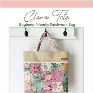 May include: A patchwork tote bag with a floral print in shades of pink, green, and yellow. The bag has a beige canvas base and a brown leather label with the text "Clara Tote" printed on it. The bag is hanging from two hooks on a white wall. The text "Molly and Mama Studio for Pretty Stitches" is printed below the bag.