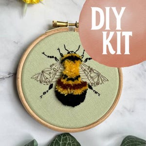 May include: A DIY kit for embroidery featuring a detailed bee design. The bee has a yellow and black body, with delicate silver wings, set against a light green fabric. The embroidery is framed in a wooden hoop, with the words "DIY KIT" in white.