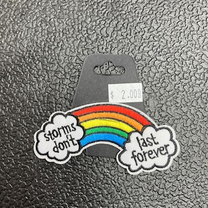 以下が含まれることがあります： 虹色のアイロン接着パッチで、白い糸で「storms don't last forever」という文字が書かれています。虹は赤、オレンジ、黄、緑、青、紫の糸で作られています。パッチは黒いテクスチャの背景にあります。