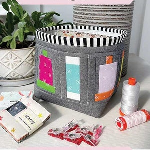 May include: A gray fabric storage basket with colorful fabric squares on the front. The basket is 6 inches wide by 5 inches high by 4 inches deep. The text "Spools Scrap Basket" is on a pink banner above the basket. The text "Finished Size 6"w x 5"h x 4"d Mini Charm and Scrap Friendly!" is on a pink banner below the basket. The text "Sew Lux Fabric" is on a white banner below the pink banner.
