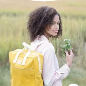 Może przedstawiać: Żółty plecak z białymi akcentami jest noszony przez osobę. Plecak ma przednią kieszeń i zamek błyskawiczny. Tekst "MAKING BACKPACK" znajduje się na górze, a pod nim "create your own backpack: regular & mini sizes".