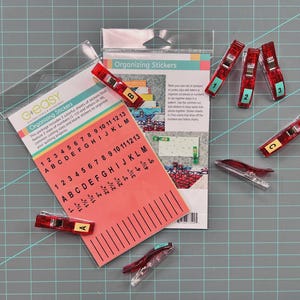 May include: A package of organizing stickers and red clips. The stickers include numbers and letters for organizing quilting projects. The red clips are labeled with numbers and letters. The package is on a gray surface.