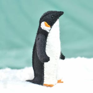 May include: A black and white felt penguin toy with orange accents. The penguin is standing on a white surface.