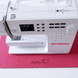May include: A white Bernina sewing machine with a digital display and control buttons. The machine sits on a pink mat with the text "Madam Sew" in cursive. The machine has a needle and thread visible.