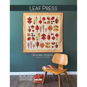 May include: A quilted wall hanging with a leaf pattern in shades of red, orange, and green, framed with a beige border. The text "LEAF PRESS" is at the top. The quilt measures 48.5 x 49.5 inches. A wooden chair sits below the quilt.