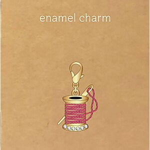 May include: Enamel charm with a gold-colored clasp and a pink thread spool design. The spool has a gold top and is adorned with clear rhinestones. The charm includes a needle and thread, and the text "JEWELRY MADE BY ME" is visible.