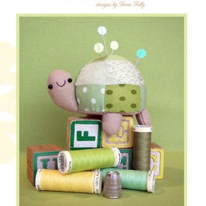 May include: A handmade turtle pincushion with a patchwork shell, adorned with pins. The image includes spools of thread, wooden blocks with letters, and a thimble. The text reads "Tiny Tim Pincushion" and "Creative Card".