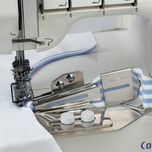Op de afbeelding: Een close-up van een naaimachine met een witte stof die door de machine wordt gevoerd. De stof wordt gevouwen over een metalen hulpstuk met een blauw-wit gestreepte band. Het hulpstuk heeft een label met de tekst "3/8 inch - 10 mm".