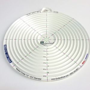 May include: A white plastic circular quilting ruler with concentric circles ranging in size from 1 1/4 inches to 6 1/4 inches in diameter. The ruler is 1/4 inch thick and has the text "Quilter's Rule" printed in the center. The text "Nested Circles 1 1/4" - 6 1/4" Diameter" is printed on the bottom edge of the ruler. The text "MQT-CT2 1/4" Thick" is printed on the left side of the ruler. The text "Made in America" is printed on the right side of the ruler.