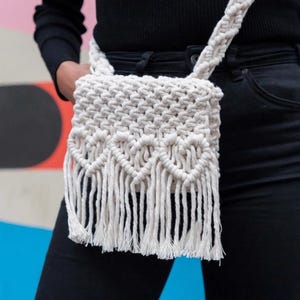 May include: A white crocheted crossbody bag with a fringe detail. The bag is worn over a black shirt and blue jeans.