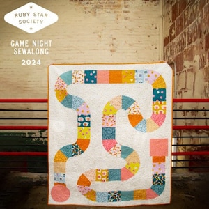 Op de afbeelding: Een wit quilt met een kleurrijk bordspelontwerp. Het quilt heeft een kronkelend pad gemaakt van vierkanten in verschillende tinten roze, blauw, geel en oranje. De tekst "GAME NIGHT SEWALONG 2024" is boven het quilt gedrukt.