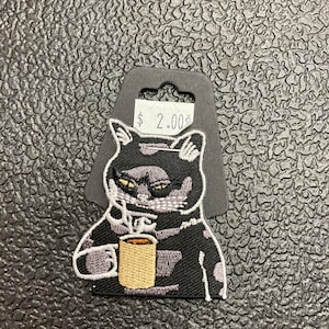 May include: Black and white embroidered iron-on patch of a cat holding a cup of coffee. The patch is on a black card with a price tag that says "$2.00".