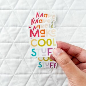 May include: A hand holding a stack of stickers with the phrase "Make COOL STUFF" in a vibrant, multi-coloured font. The stickers are against a white, quilted background. The text uses a mix of colours, including red, orange, yellow, green, purple, and blue.