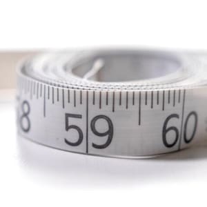 Op de afbeelding: Een opgerold wit en grijs meetlint met de cijfers 58, 59 en 60 duidelijk zichtbaar. De tape is gemarkeerd met zwarte stappen en cijfers, die inches aangeven. De equivalent van 152 cm is zichtbaar. De achtergrond is wit.