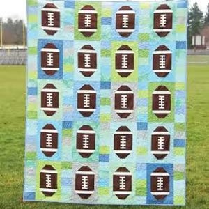 May include: A handmade quilt featuring a repeating pattern of brown footballs on a patchwork background of blue, green, and white squares. The word "Touchdown" is at the top. The quilt is displayed on a grassy field.