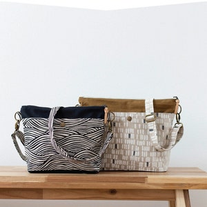 May include: Two tote bags, one black and white with a wave pattern and one beige with a white and gray geometric pattern, are displayed on a wooden table. The bags have brown leather straps and are labeled "Hillside Tote" and "noodlehead".