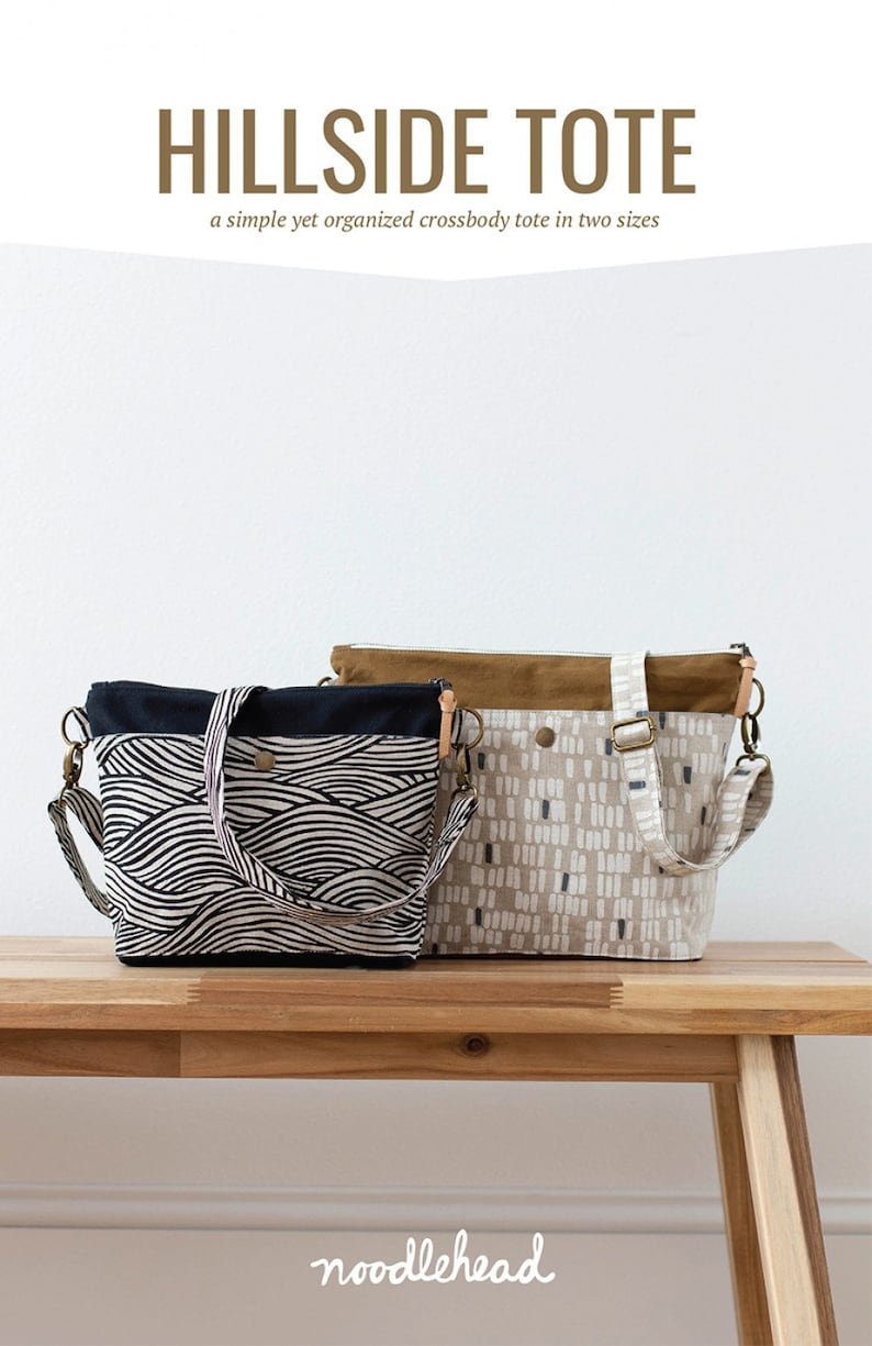 May include: Two tote bags, one black and white with a wave pattern and one beige with a white and gray geometric pattern, are displayed on a wooden table. The bags have brown leather straps and are labeled "Hillside Tote" and "noodlehead".