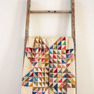 May include: A patchwork quilt with a star design, displayed over a wooden ladder. The quilt has a cream-colored base with gold polka dots and a star pattern composed of colourful triangles in various patterns and shades, including red, blue, and orange.