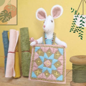 May include: A handcrafted white felt mouse figurine wearing blue overalls and holding a small, square, quilted fabric piece featuring a geometric star pattern in pink, blue, and olive green, accompanied by felt fabric rolls and a wooden spool of thread.