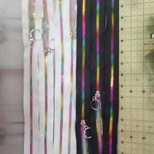 May include: A collection of zipper tapes featuring rainbow-colored teeth on white and black fabric strips. Each zipper includes a circular metal pull tab. A yellow measuring ruler is positioned on the right, showing a length of 8 inches.
