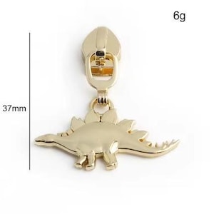 May include: Gold-toned zipper pull featuring a 37mm dinosaur charm. The dinosaur is a stegosaurus with a spiky back and a long tail. The zipper pull weighs 6 grams.
