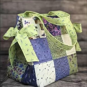 May include: A patchwork tote bag with a light green handle and bow. The bag features a variety of fabric patterns, including floral, botanical, and abstract designs in shades of green, blue, and white. The bag is sitting on a wooden surface.