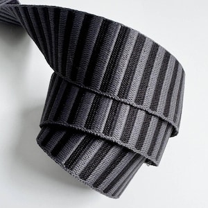 Puede incluir: Una longitud enrollada de cinta elástica tejida que presenta un patrón repetitivo de rayas verticales en tonos alternos de gris carbón y negro. La tela tiene una textura acanalada distintiva con un ancho constante y una construcción flexible.
