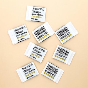 May include: White fabric labels with black text that reads "Beautiful Things come together One Stitch at a time." and "HANDMADE WITH" with heart icons. The labels are scattered on a light beige surface.
