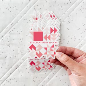 May include: A stack of stickers with a geometric quilt block design in shades of pink and white. The stickers have the text "I STILL PLAY WITH BLOCKS" printed on them. The stickers are held by a hand.