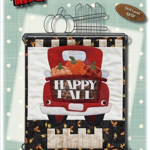 Op de afbeelding: Een rode vrachtwagen met een wit "Happy Fall"-bord aan de achterkant, gevuld met oranje pompoenen. De vrachtwagen staat op een zwart-wit geblokte achtergrond met herfstbladeren. De afbeelding is voor een quiltpatroon.