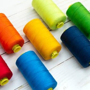 Pode incluir: Uma coleção de carretéis de linha coloridos, incluindo vermelho, laranja, amarelo, verde, azul e roxo. Cada carretel tem uma base amarela. As linhas estão dispostas sobre uma superfície de madeira clara.