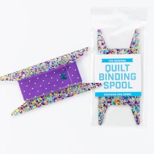 Puede incluir: Un carrete de plástico cubierto de purpurina de colores, con forma de cabeza de gato con dos orejas puntiagudas, está envuelto en una tela de lunares blancos y morados. El embalaje dice "The Original Quilt Binding Spool" y "Contains One Spool".