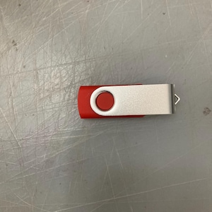 May include: A red and silver USB flash drive. The red portion has a circular button and the silver portion has a small loop. The flash drive is lying on a gray surface.
