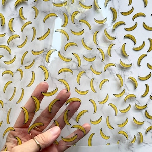 May include: A clear acrylic sheet featuring a repeating pattern of small, yellow banana illustrations. The sheet is transparent, allowing the underlying surface to be visible through the material between the printed banana graphics.