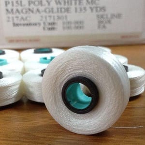 May include: Close-up of white thread spools with black and teal centers. The spools are arranged on a wooden surface. The text on the box reads "FIL-TEC" and "MAGNA-GLIDE 135 YDS".