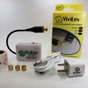 May include: A white rechargeable laser system with a green laser, a white power adapter, a white USB cable, three gold-coloured laser heads, and a roll of white stickers with red markings. The box for the laser system is yellow with the text "ViviLux 3-in-1 Rechargeable Laser System CrossHair Line Dot No Batteries Needed!" The box also has a picture of the laser system being used on a surface.