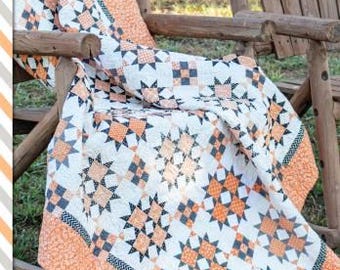 Pode incluir: Um cobertor com tema de Halloween com estrelas e quadrados laranja, preto e branco. O cobertor está drapeado sobre uma cadeira de madeira com o texto "Halloween Memories" acima. O cobertor é de Susan Ache.