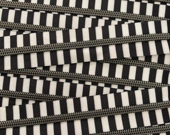 May include: Close-up of black and white striped zipper tape. The tape is made of a woven fabric with a silver metal zipper.