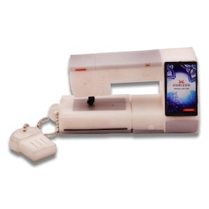 May include: A white and gray miniature sewing machine with a keychain attached. The machine has the brand name "Janome" printed on it, along with the model name "Horizon Memory Craft 15000". The keychain is shaped like a sewing machine.
