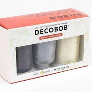 May include: A rectangular white and red box containing three spools of Decobob cottonized polyester thread. The packaging displays the text Decobob, 3 Spools, 2000m (2187yd), and Violet Craft x Wonderfil. The spools are dark gray, light gray, and cream.