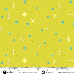 May include: A fabric with a bright yellow background featuring a repeating pattern of white starbursts, small brown dots, and turquoise star shapes. The fabric is marked with a ruler along the bottom edge.