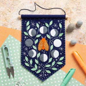 Op de afbeelding: Een marineblauwe vilten wandbanner met een centrale oranje mot omringd door geborduurde lichtgroene bladeren, witte maanfasen en kleine sterren. De banner hangt aan een houten stok met een zwart koord en heeft witte stiksels.