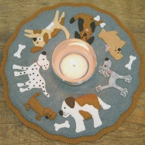Puede incluir: Un tapete de fieltro para velas con un fondo azul claro y un borde festoneado. El tapete presenta figuras de perros aplicadas y formas de huesos blancos. Un portavelas de vidrio transparente con una vela encendida se encuentra en el centro.